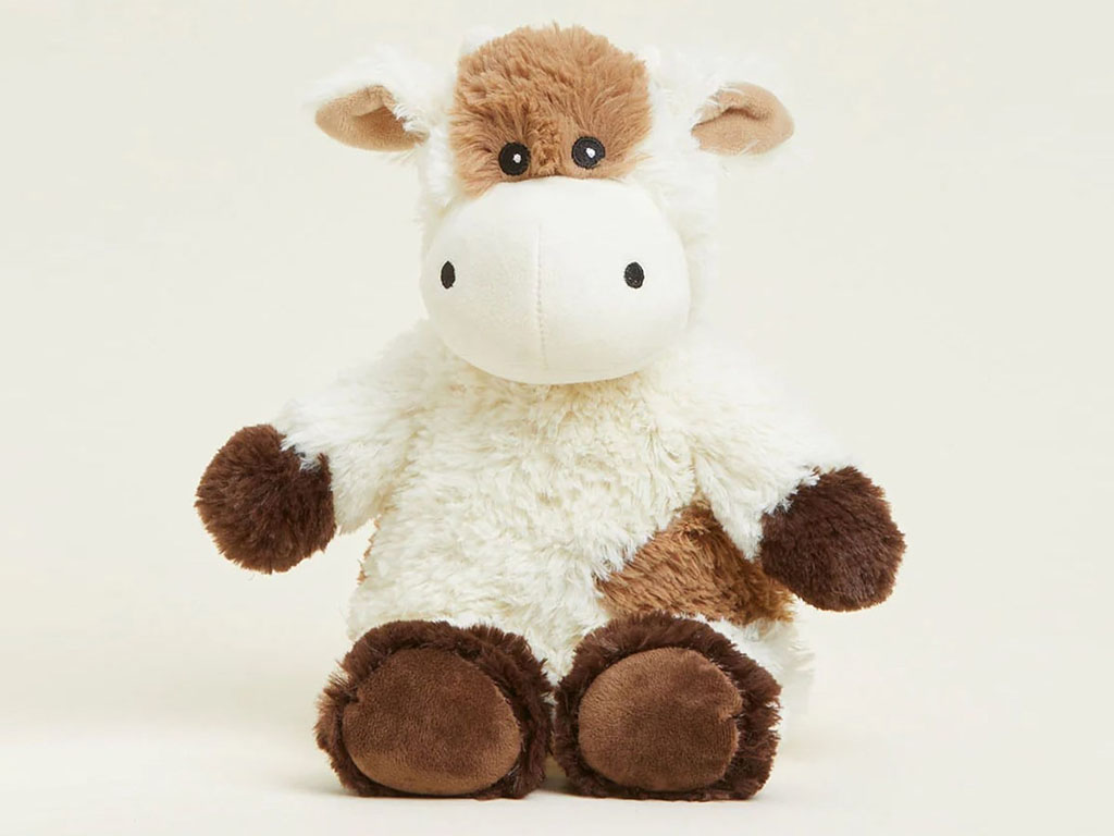 A soft plush cow toy sitting upright against a light background, featuring a white fluffy body, brown patches, rounded hooves, small horns, and a friendly embroidered face.