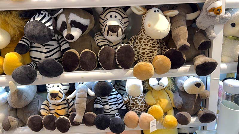 Retail display of assorted zoo animal plush toys including zebra, giraffe, elephant, raccoon, and bear designs, arranged on white shelves, featuring soft plush fabrics, neutral color palettes, and child-safe construction suitable for gift shops, toy stores, and wholesale or OEM plush toy sourcing.
