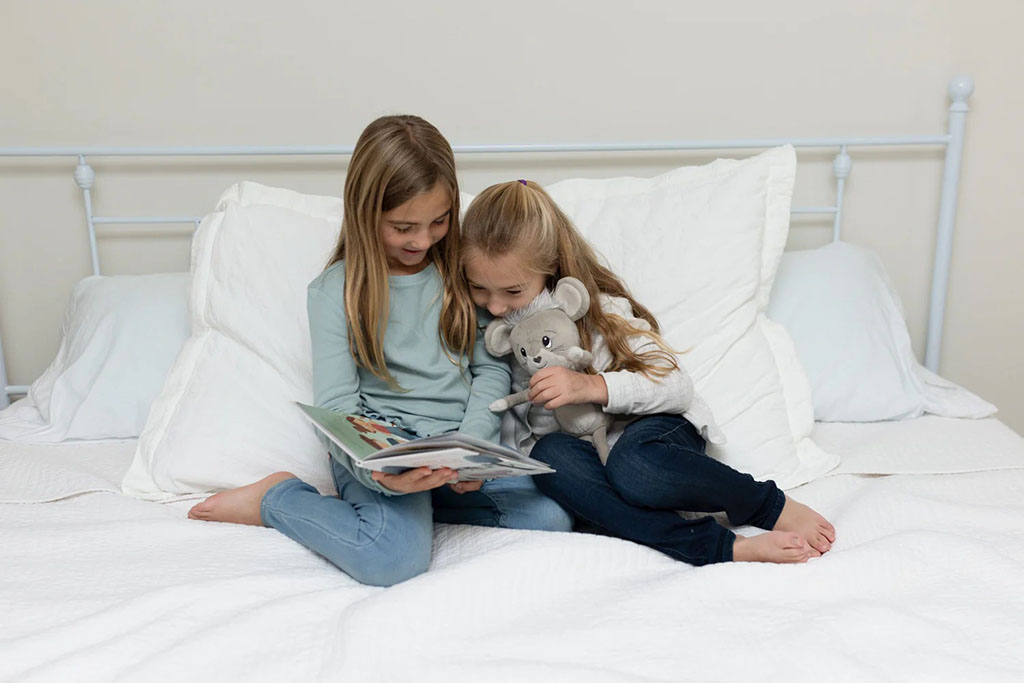 Two young girls sitting together on a white bed, reading a picture book while cuddling a soft gray stuffed animal, creating a calm and comforting bedtime moment
