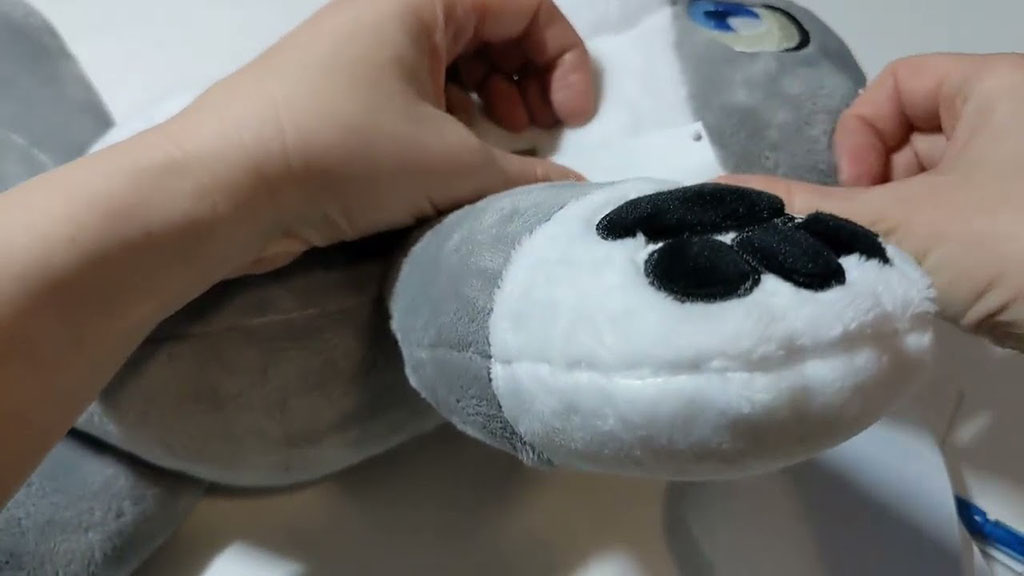 Hands fixing or adjusting a grey and white plush animal, focusing on its paw and stitching area.
