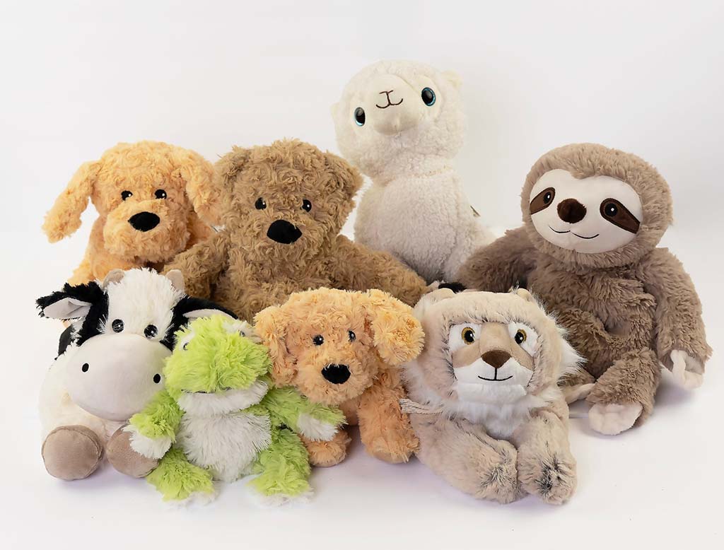 Cute grey sloth plush toy sitting on a rustic wooden surface with decorative greenery and books in the background. The sloth has a friendly embroidered face, long limbs, and fluffy faux-fur fabric, making it ideal for kids or collectors.