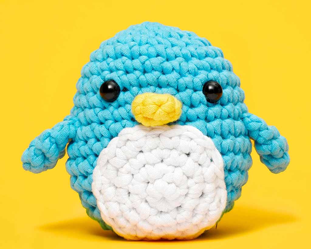 Handmade crocheted penguin plush toy with blue and white yarn texture, featuring stitched details and a rounded amigurumi design, displayed on a yellow background to highlight soft yarn materials, artisanal craftsmanship, and inspiration for custom handmade plush toy production.