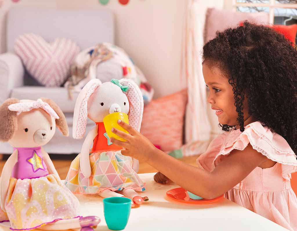 A young girl sits at a table smiling as she pretends to serve a drink to two plush animal dolls dressed in colorful outfits during imaginative playtime.