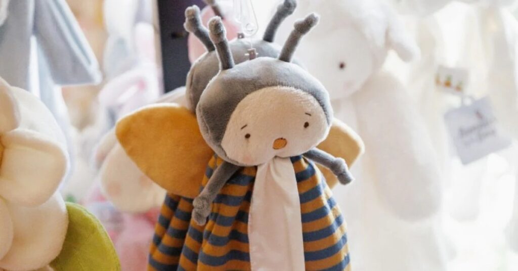 Soft bee-shaped plush toy hanging in a baby boutique display, featuring gentle pastel colors, embroidered facial details, and lightweight construction, ideal for illustrating baby-safe stuffed animal design, sensory comfort toys, and premium plush craftsmanship for infant and newborn product collections.