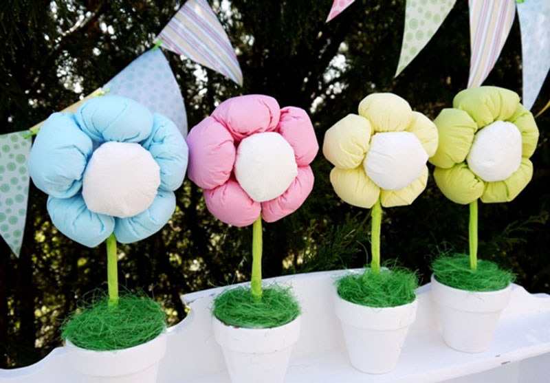 Four colorful plush flowers in pastel blue, pink, yellow, and green displayed in white pots with green grass-like filler at an outdoor setting.