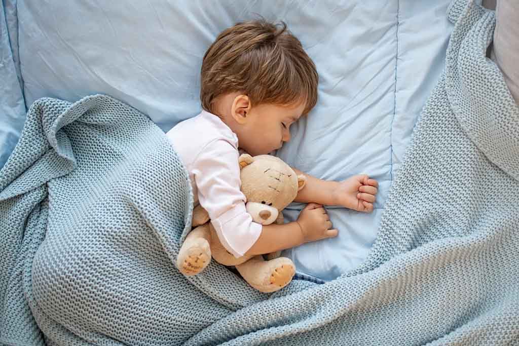 A toddler sleeps peacefully on a light blue bed while hugging a soft beige teddy bear, wrapped partly in a knitted blue blanket.
