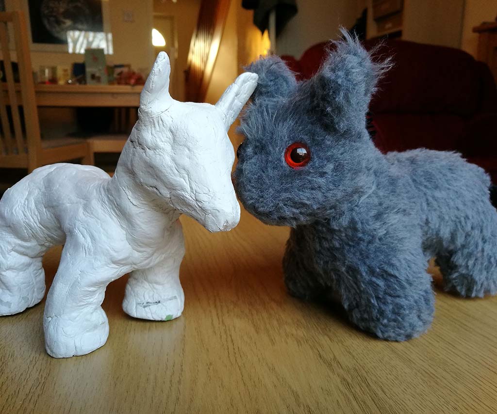A gray plush animal with bright red eyes facing a white handmade clay animal figure on a wooden table, creating a playful face-to-face contrast between soft toy and sculpted figure