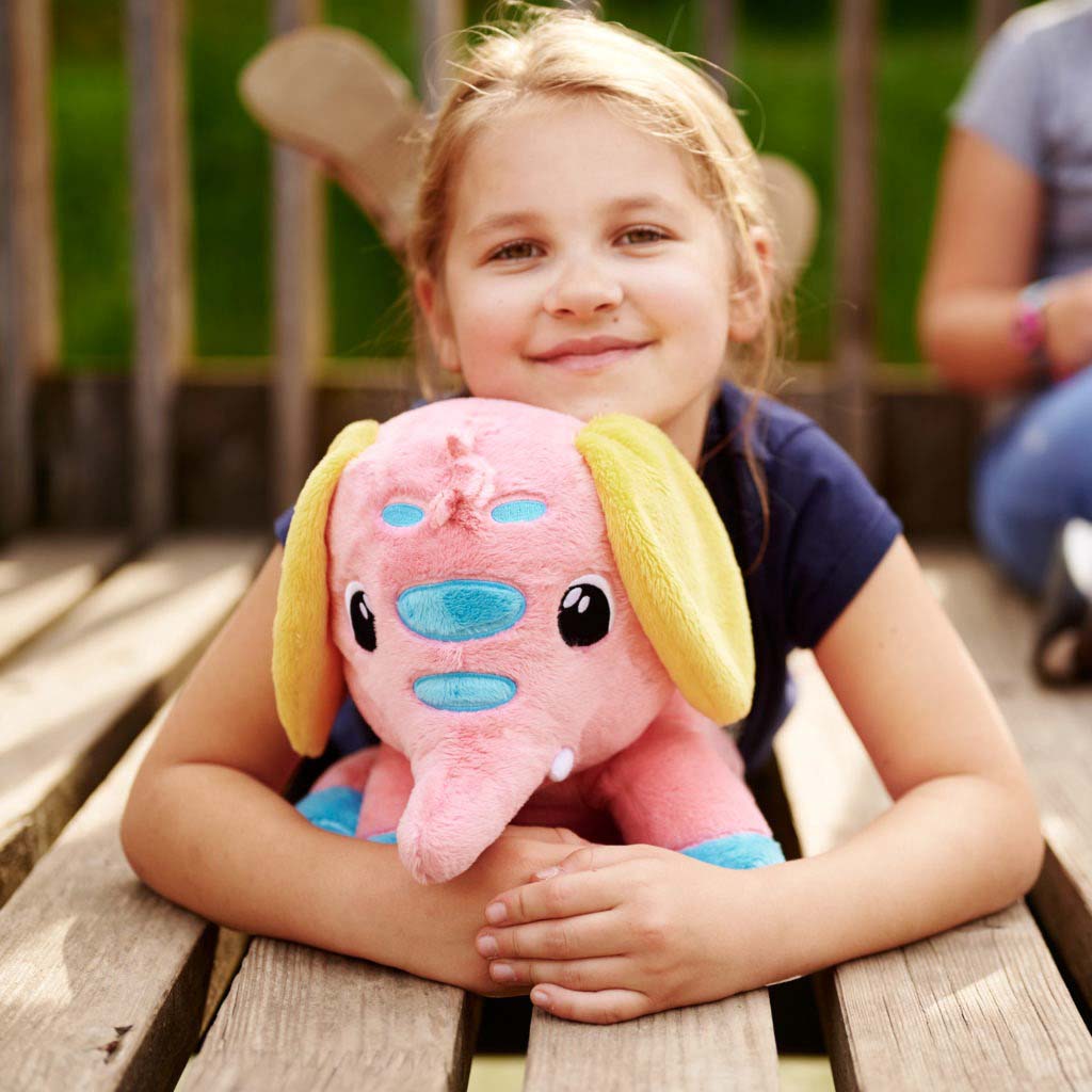 A smiling young girl outdoors hugging a pink elephant plush toy with yellow ears and blue facial details while sitting on wooden steps.