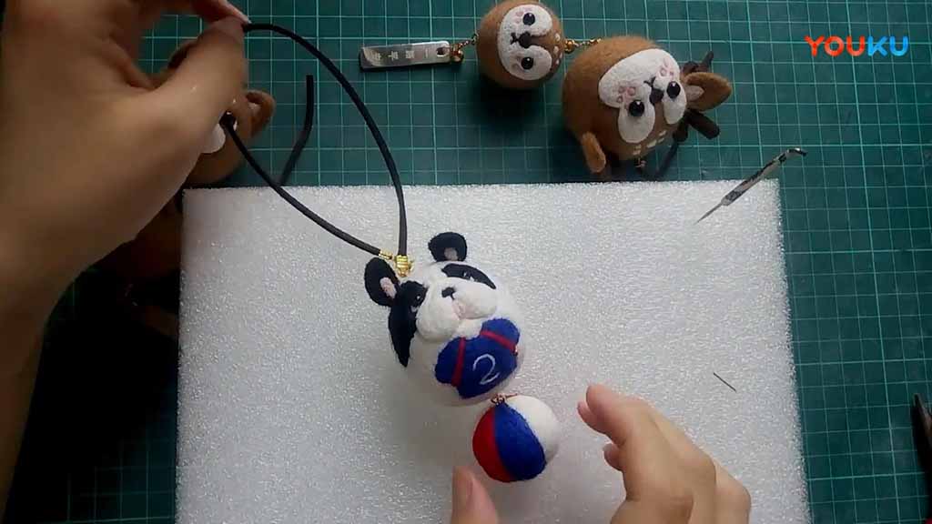 Hands assembling a handmade panda plush keychain on a foam board, with small felt animal keychains and crafting tools placed around on a green cutting mat.