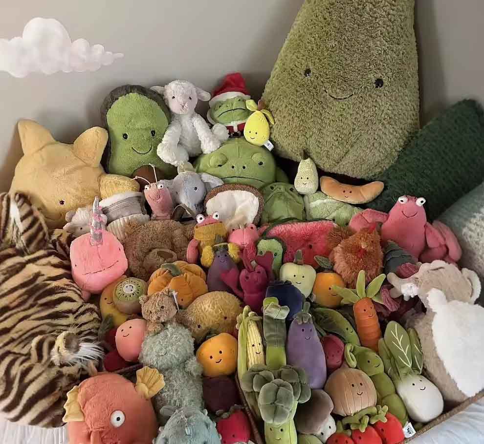 Large assorted collection of Jellycat plush fruits, vegetables, animals, and characters arranged together, including broccoli, peas, avocado, carrot, eggplant, berries, and whimsical creatures in a cozy display.