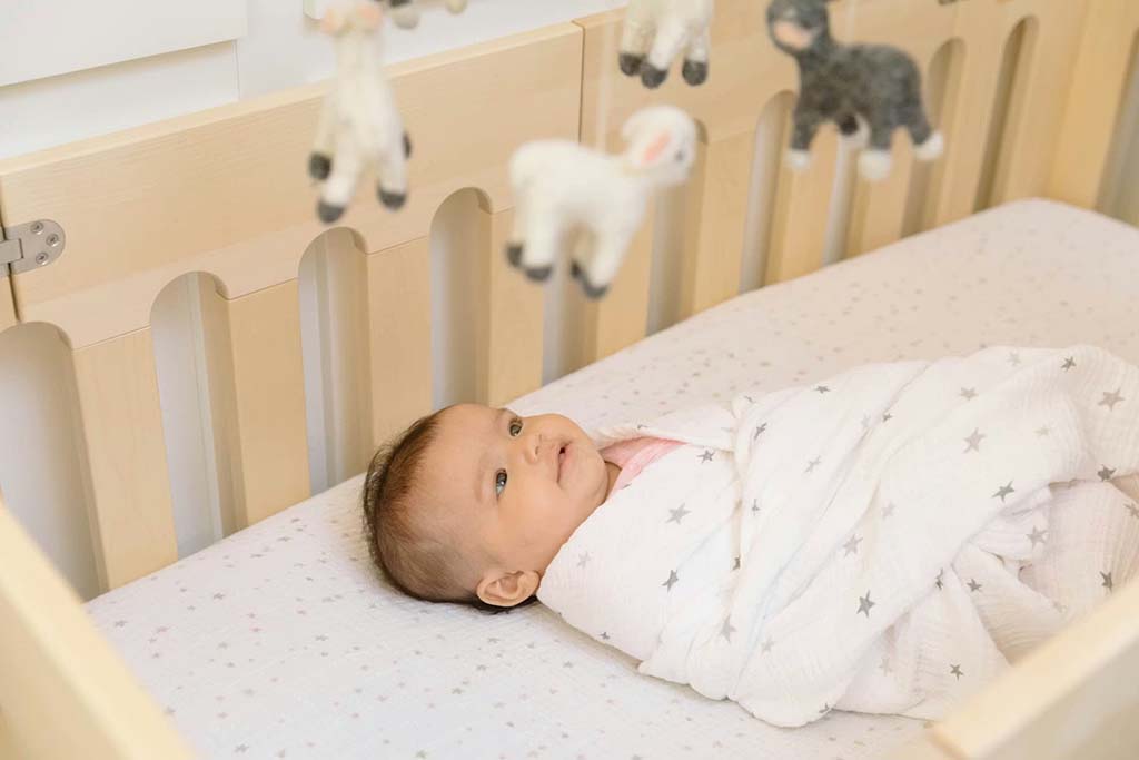 A swaddled infant lies safely in a wooden crib, looking up at a soft animal mobile hanging above.