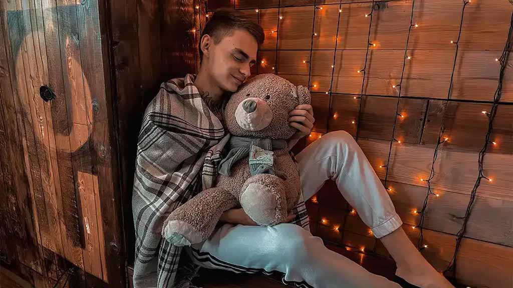 A young man sits wrapped in a blanket, peacefully cuddling a teddy bear against a backdrop of warm string lights, illustrating the calming and emotional comfort adults find in plush toys.