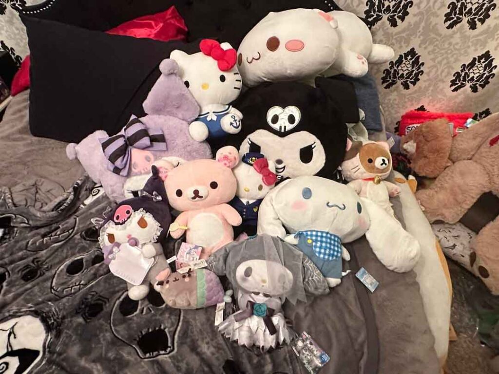 A large collection of cute plush toys, including Sanrio characters like Hello Kitty, Kuromi, My Melody, Cinnamoroll, and other soft animals, arranged together on a bed with decorative blankets and pillows.