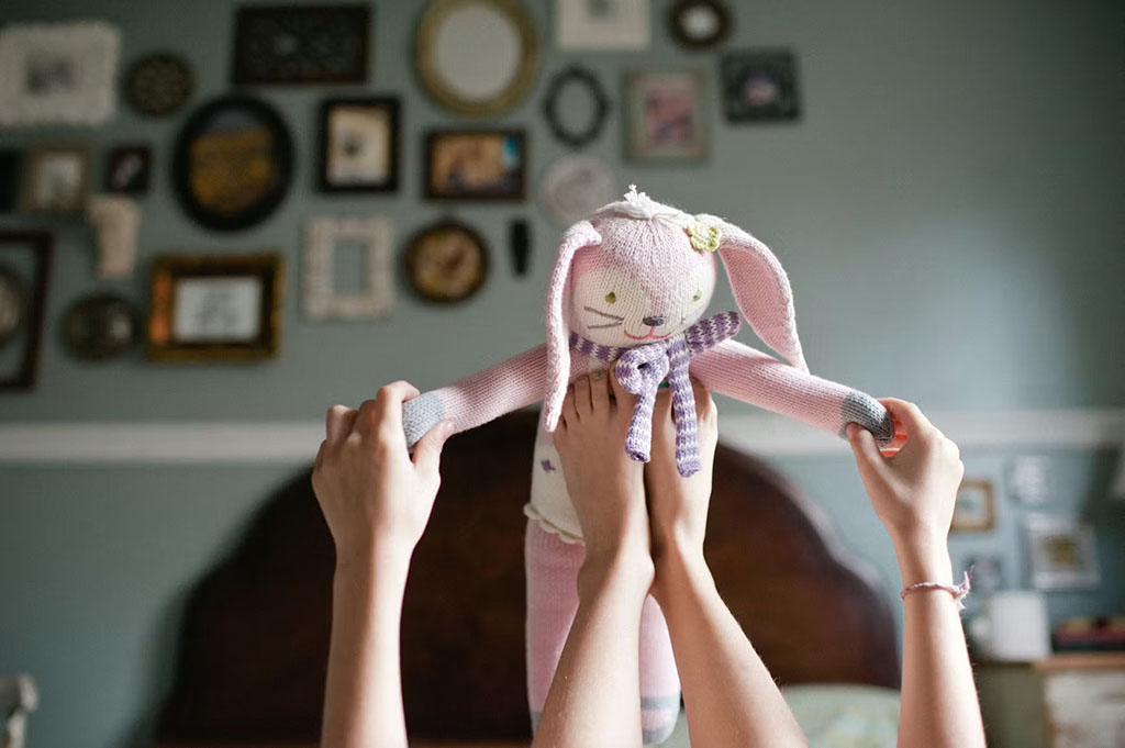 A child is holding a knitted bunny plush by its arms, with their feet raised and the plush dangling in between. The background features a room decorated with various photo frames on the wall.