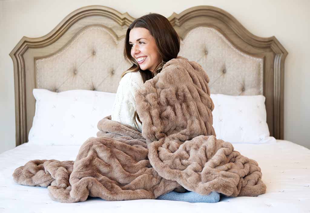 Woman wrapped in thick plush minky blanket while sitting on bed, highlighting ultra-soft texture, high-pile fabric surface, draped weight, and premium comfort design, ideal for custom minky blanket manufacturing, private label home textiles, gift blankets, and high-end plush fabric bedding collections.