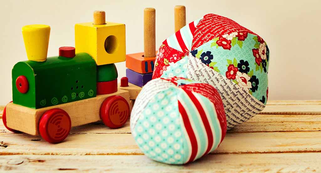 Colorful handmade fabric plush balls with patchwork cotton panels placed beside a wooden toy train on a light wooden surface, showing soft baby-safe texture and playful patterns.