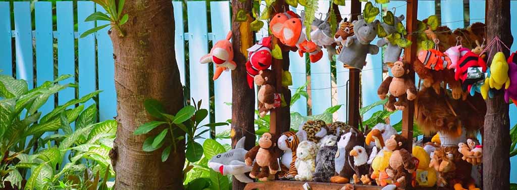 A vibrant outdoor market-style display of assorted plush toys hanging and arranged on wooden beams, featuring animals like monkeys, sharks, birds, and tigers with green plants and a blue fence in the background.
