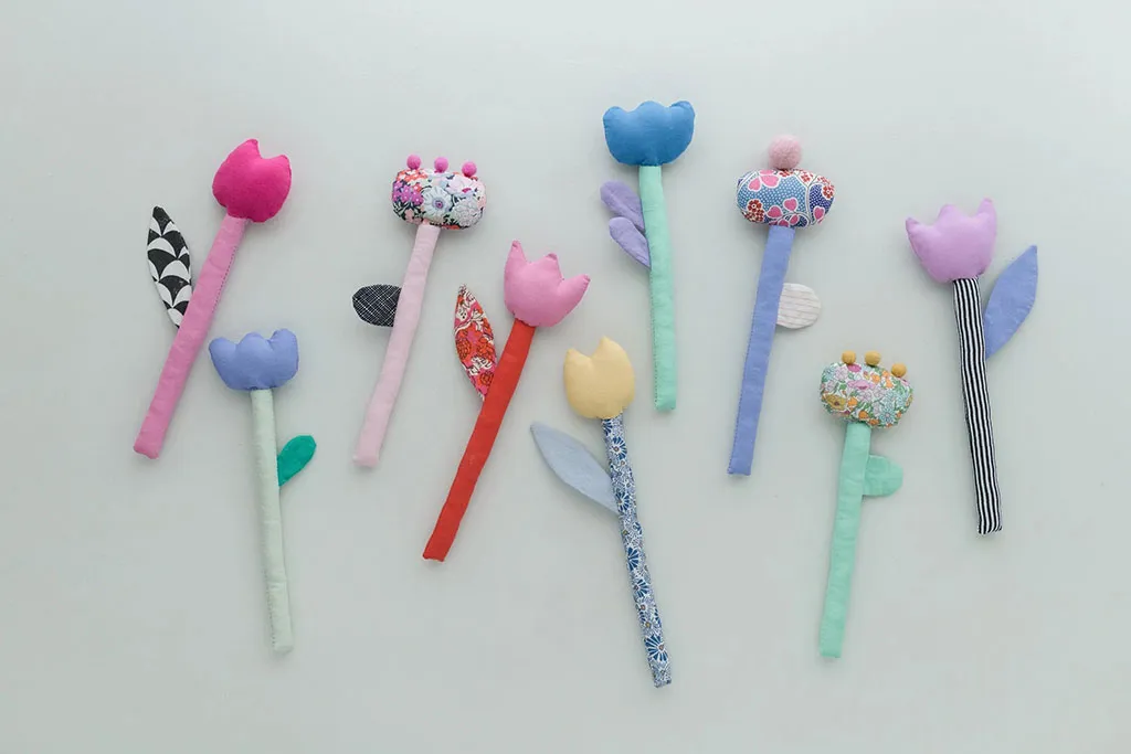 Handmade plush flower stems in various colors and patterns arranged on a light background, featuring soft fabric petals, leaves, and decorative details.