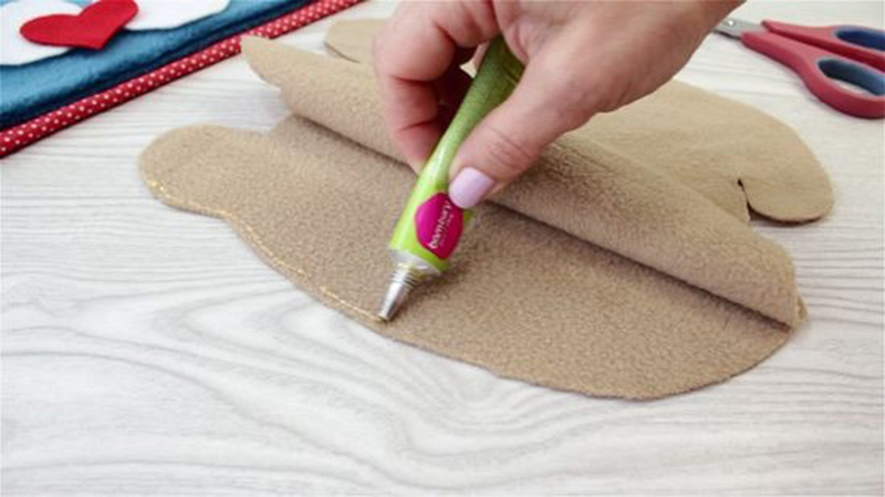 A hand applying fabric glue along the edge of beige plush material pieces while crafting a stuffed animal, with scissors and sewing supplies in the background.