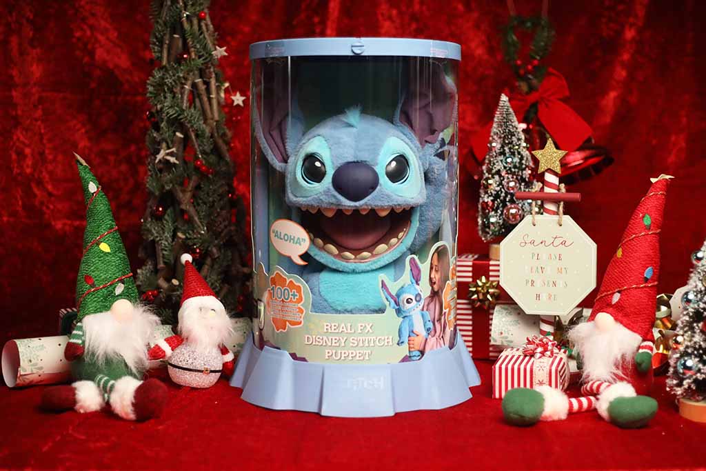 A boxed Disney Stitch Real FX puppet displayed in a festive Christmas scene with red velvet background, small decorative gnomes, miniature trees, wrapped gifts, and a holiday sign, creating a cheerful holiday atmosphere.