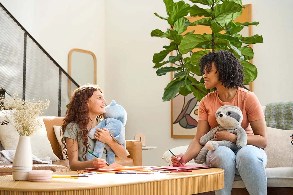 Mother and young girl sitting together at a coffee table, drawing with colored pencils while each holds a soft plush sloth toy in a bright, modern living room.
