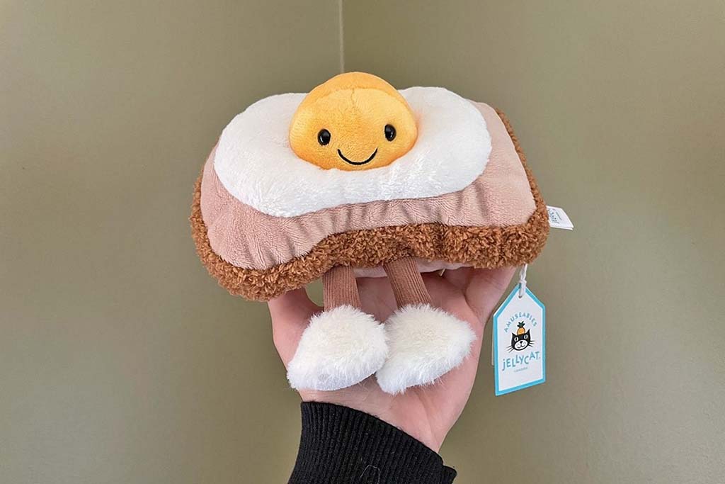 A hand holding a Jellycat plush shaped like toast with a smiling fried egg on top and fluffy dangling legs.