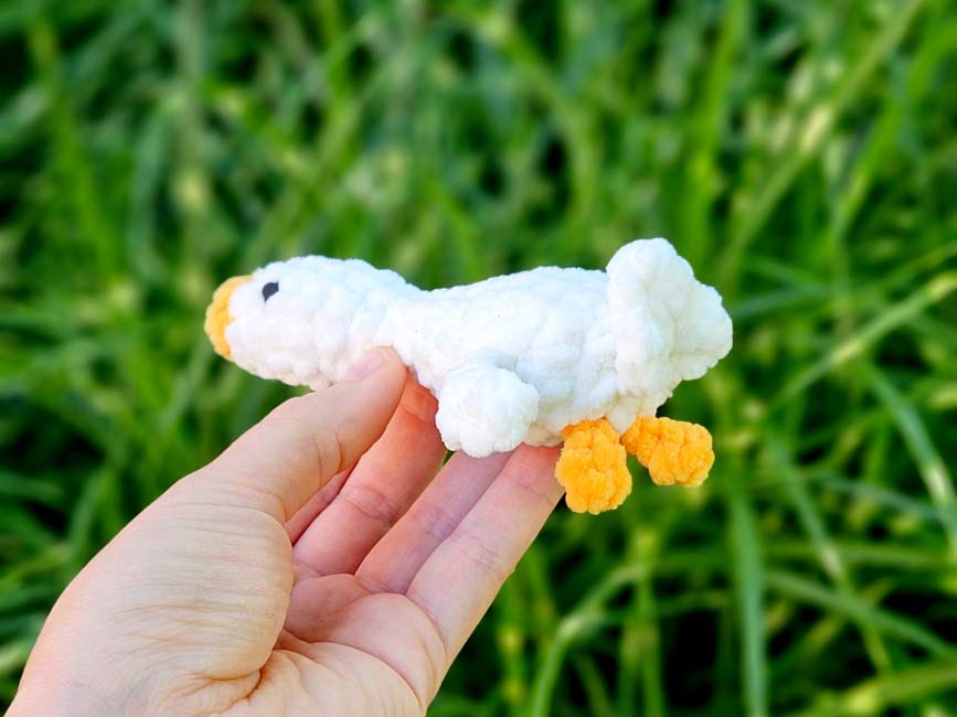 Small handmade plush duck with white textured body, yellow beak, and orange feet, held in a hand outdoors against green grass background.