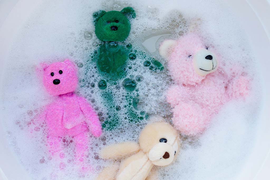 Four small plush toys soaking in soapy water inside a bathtub, including a pink bear, green bear, cream-colored bunny, and pink teddy, illustrating the process of washing stuffed animals.