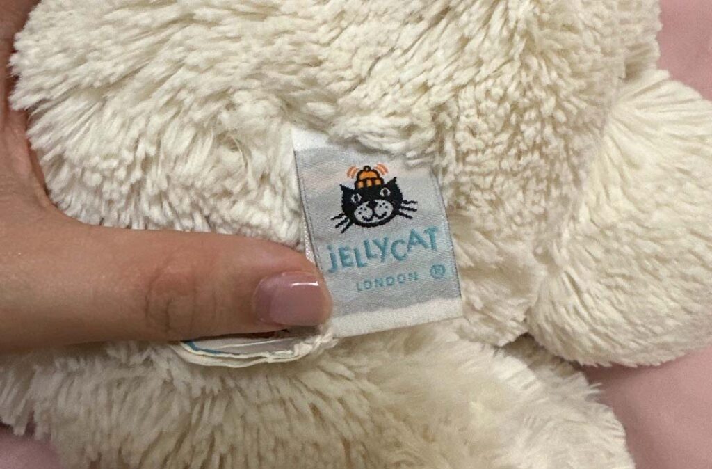 Close-up photo of a Jellycat London plush toy tag being held by a hand, showing the classic Jellycat cat logo and soft cream-colored fur of the stuffed animal.