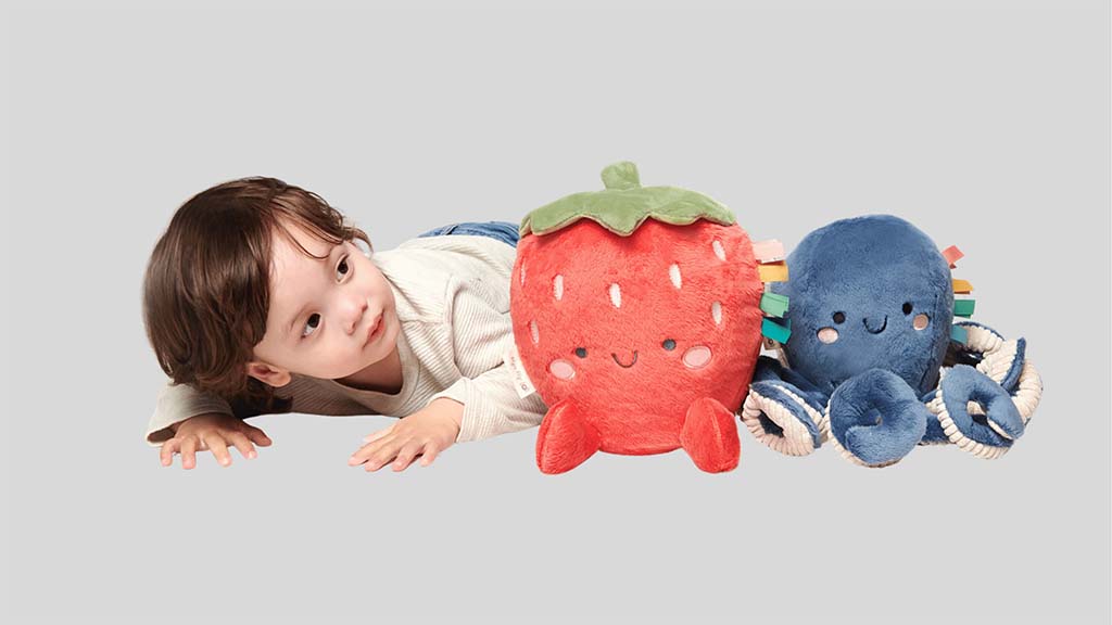 Baby lying on the floor and reaching forward beside two soft plush toys—a smiling strawberry plush and a blue octopus plush—designed for sensory play and early development.