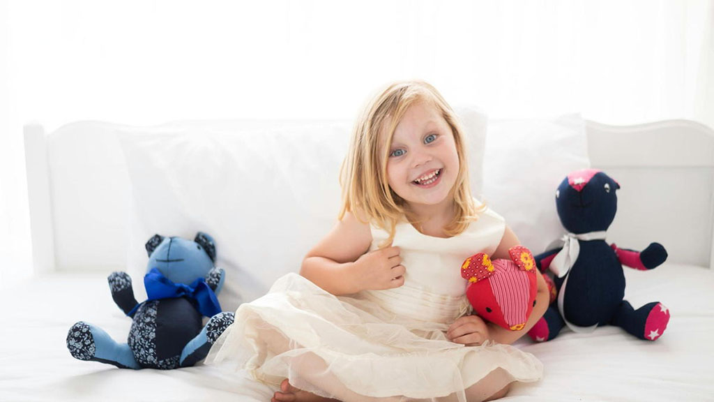A smiling young girl in a white dress sits on a bed, holding a bright pink plush toy. Two other colorful plush bears are positioned next to her, one in blue with a bowtie and the other in dark blue with red and white stars. The background features soft white curtains.