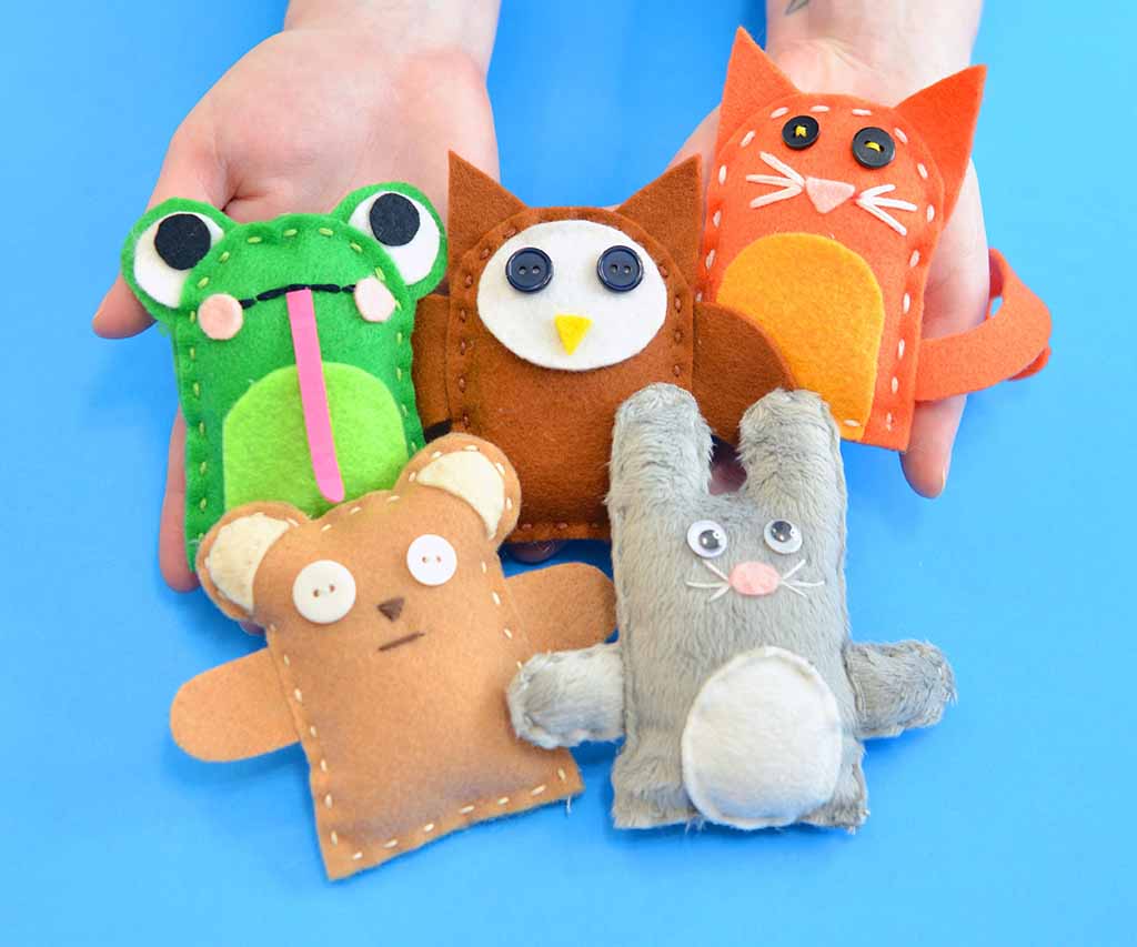 Five small handmade felt plush animals—a frog, owl, cat, bear, and bunny—displayed in two hands against a blue background.