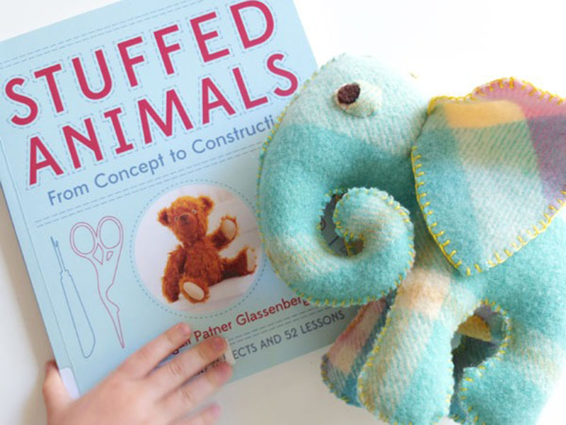A child’s hand holds a stuffed-animal crafting book beside a handmade teal and plaid felt elephant plush with yellow stitching.