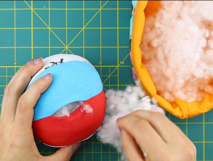 Hands stuffing polyester fiberfill into a colorful fabric plush ball through an opening, showing the soft filling material and sewing workspace on a cutting mat.