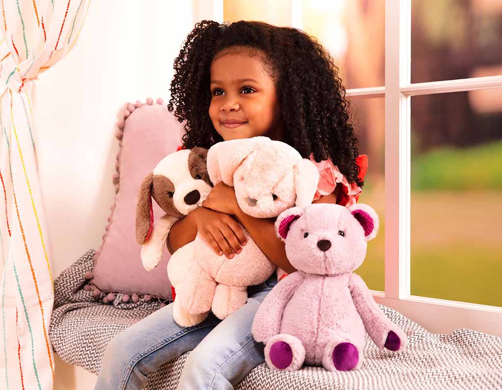 A young girl sits by a window smiling and hugging soft plush toys, including a stuffed dog and teddy bears, in a cozy, brightly lit room.