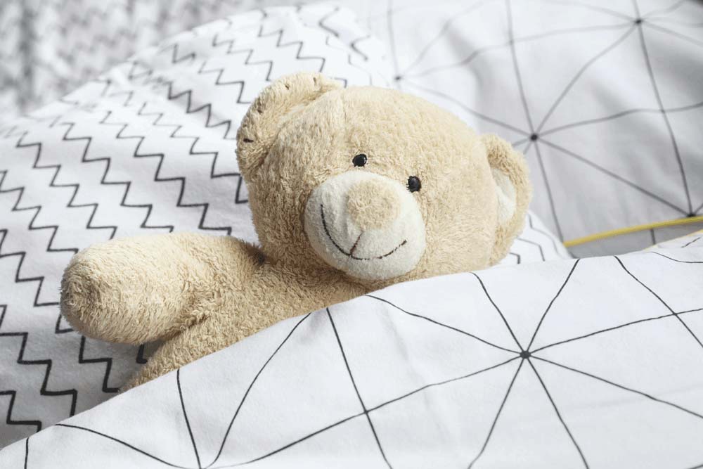 A soft beige teddy bear tucked under white patterned bedding, lying on a bed with geometric black-line designs, creating a cozy and comforting scene.