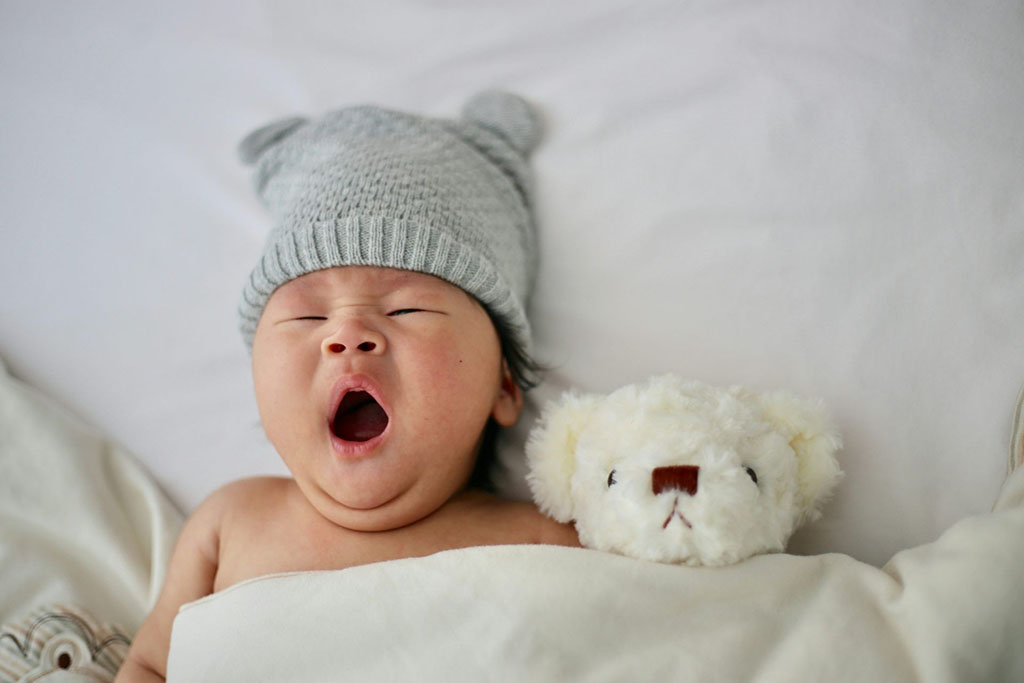 Baby yawning in bed with a soft teddy bear and knit hat.