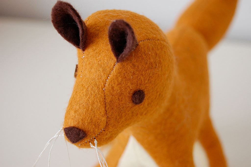 A close-up of a handmade felt fox plush toy with stitched details, brown ears, and white whiskers.