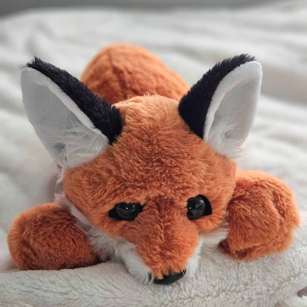 A soft orange fox plush toy lies on a white blanket, featuring large pointed ears, fluffy fur, and realistic black eyes, creating a cute and comforting appearance.