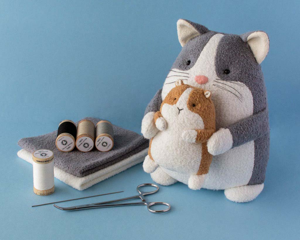 Handmade grey and white cat plush holding a small brown and white hamster plush, displayed with sewing tools, fabric pieces, and thread spools on a blue background.