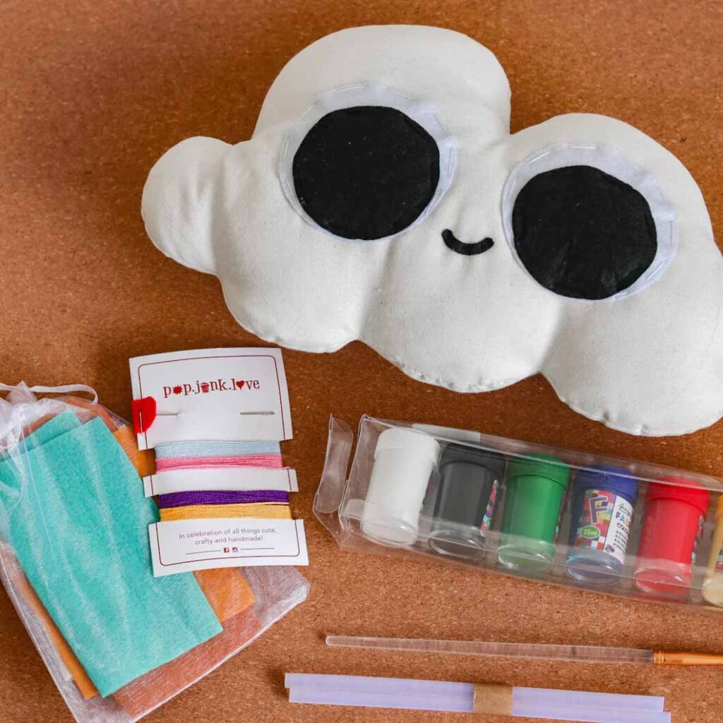 A handmade cloud-shaped plush with large black eyes, surrounded by crafting supplies including felt pieces, thread, paints, and tools.