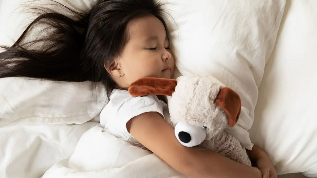 A young girl sleeps peacefully in a white bed while hugging a soft beige puppy plush toy with brown ears, conveying comfort, security, and warmth.