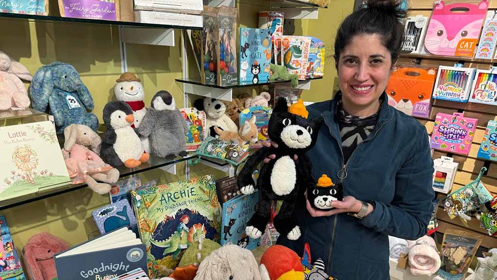 Woman smiling inside a gift and toy store while holding two soft plush toys, surrounded by stuffed animals and children’s books on retail shelves, representing plush toy merchandising, gift shop displays, and specialty toy retail environments.