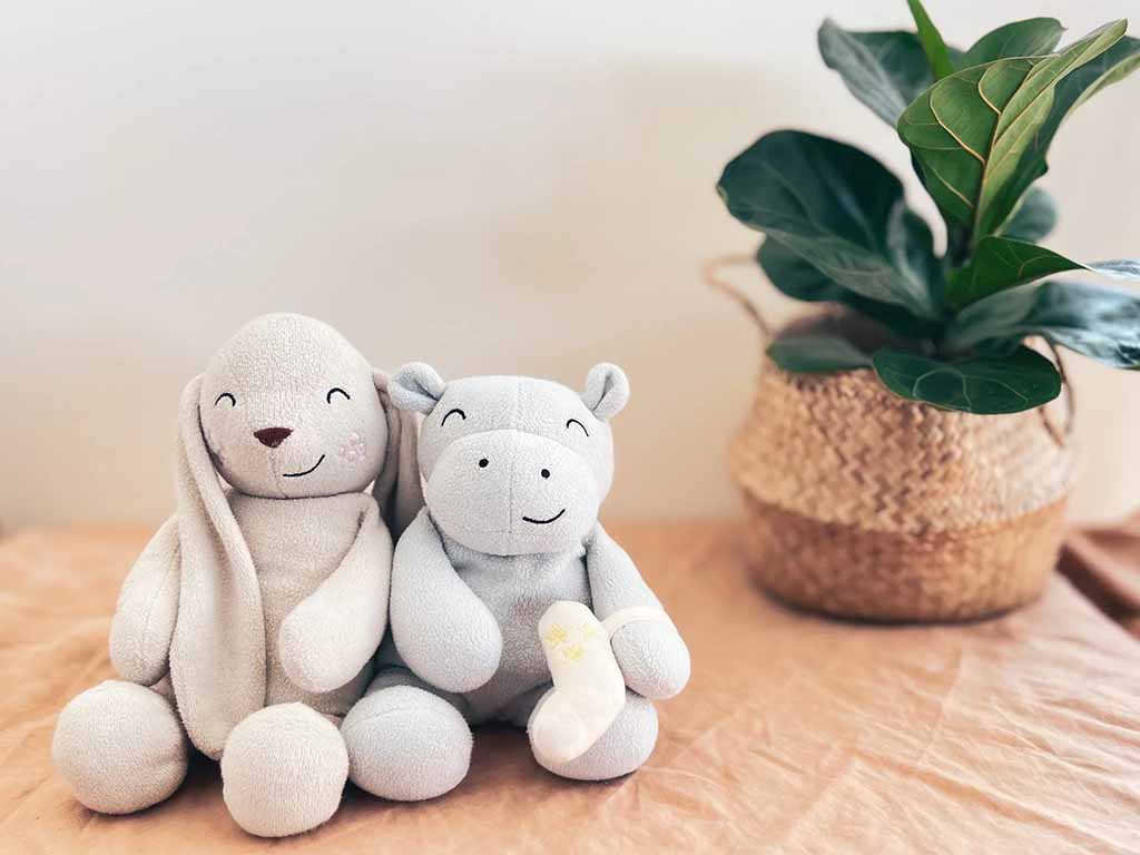 A soft, light gray plush bunny and bear sitting side by side on a table, with a potted plant in the background. The bunny has long ears and a smiling expression, while the bear holds a small toy.