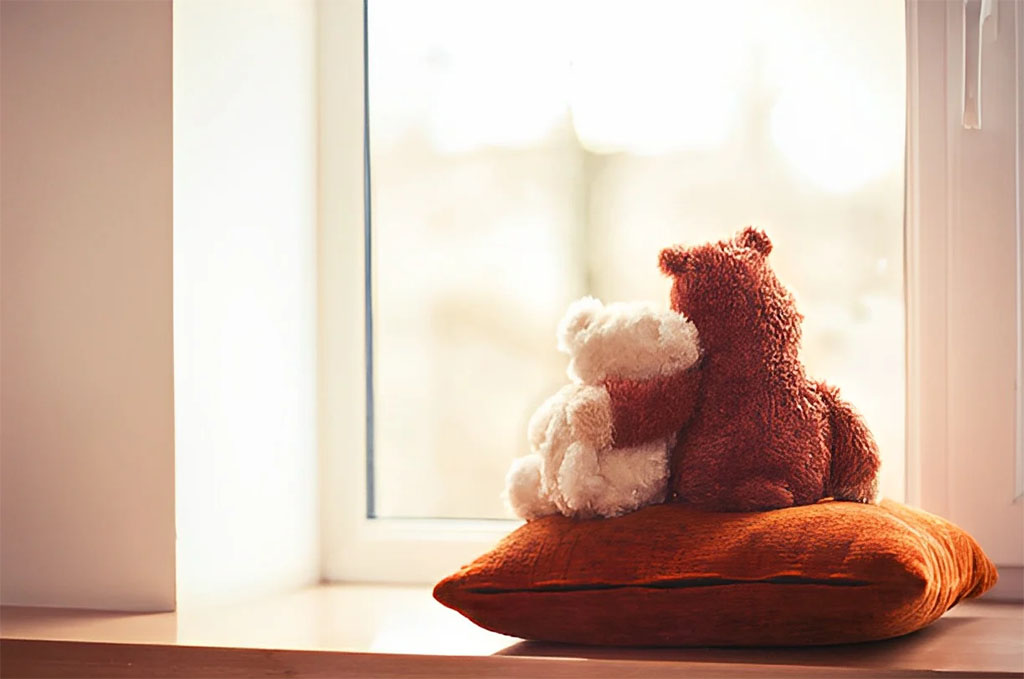Two teddy bears hugging while sitting on a cushion by the window.