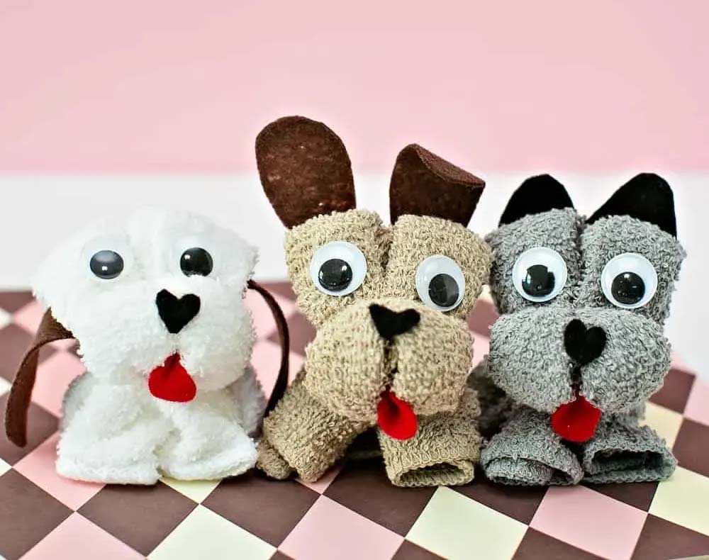 Three handmade towel puppies with googly eyes, felt ears, and red felt tongues displayed on a pink and brown checkered surface.