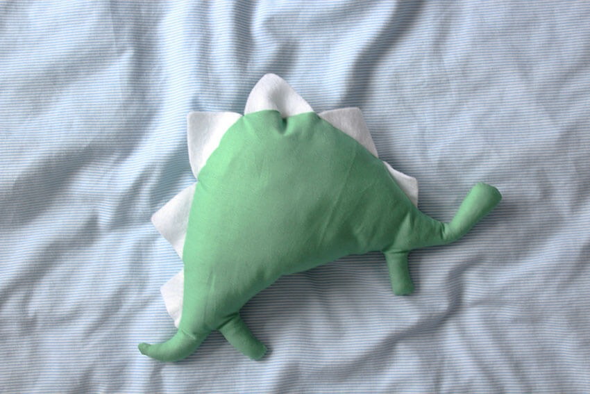 A simple handmade green dinosaur plush toy with white felt spikes, lying flat on a light blue fabric background, showing the completed sewing and stuffing result.