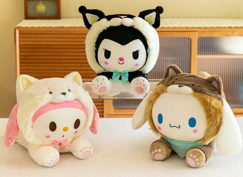 Three round plush toys wearing soft animal hood costumes, including a black character with a teal bow, a pink-themed bunny plush, and a blue-cheeked plush in a brown animal hood, displayed on a table.