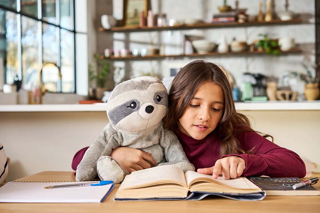 Young girl reading a book at a table while hugging a soft sloth plush toy, with notebooks and pencils nearby in a cozy home setting.