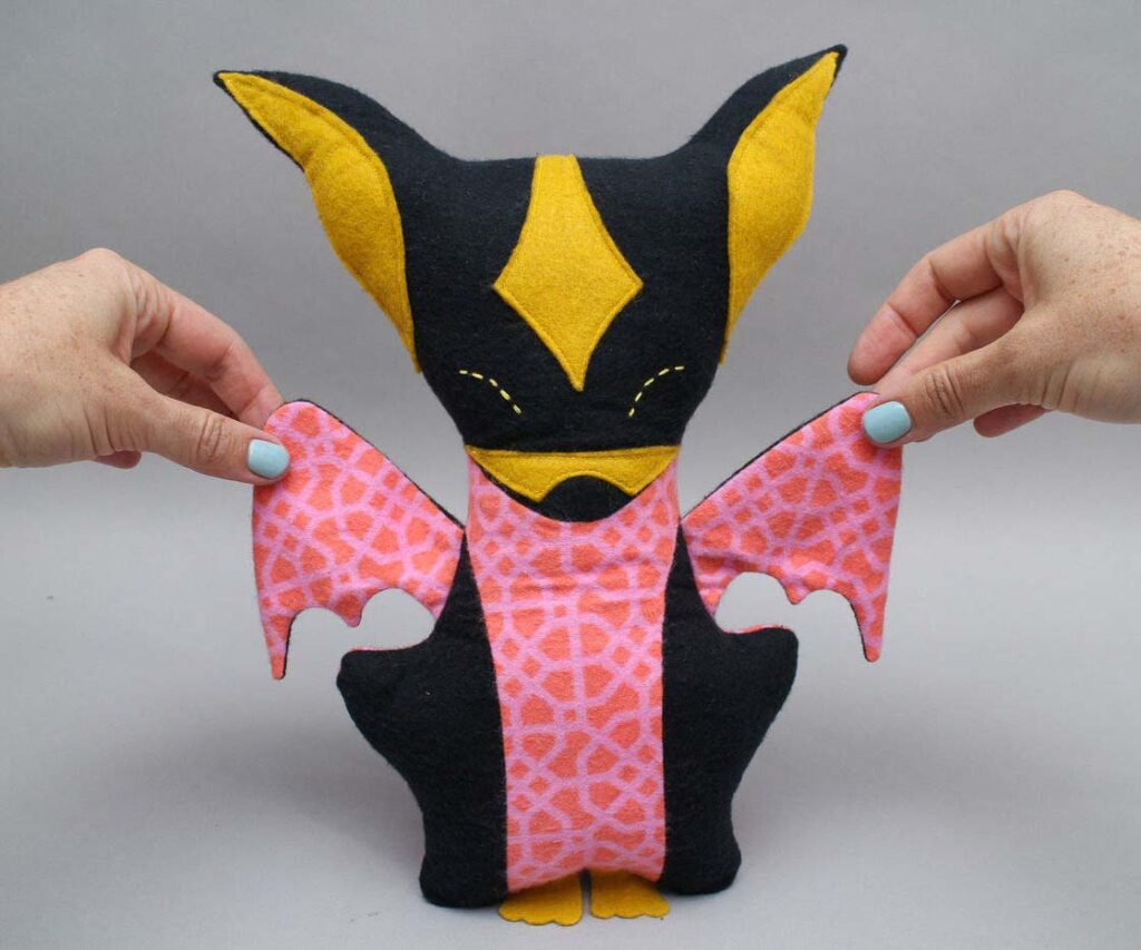 Handmade bat plush toy crafted from black, yellow, and patterned pink felt, with two hands holding its wings open to display the shape. The plush features tall ears, stitched facial details, and decorative geometric pink wings.